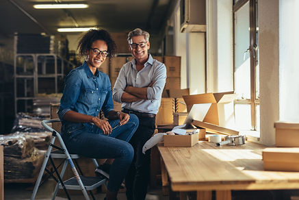 Woman and man, business owners, happily sitting in their shipment room. They are an example of the type of client that can benefit from personal wealth management.