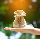 A flat hand holding a bag with a dollar sign printed on it, as a symbol of obtaining money through life events like sale of business, inheritance, and real estate sales, as IronBridge Private Wealth can help through these events.
